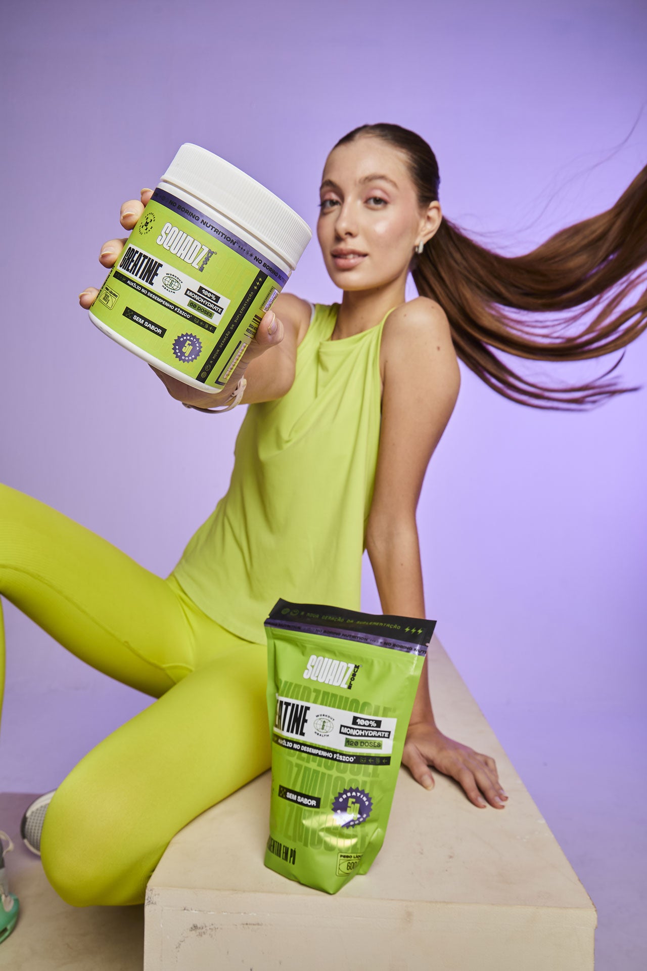 Mulher de macacão verde-limão sentada ao lado do pote e do refil de Creatine SQUADZ MUSCLE