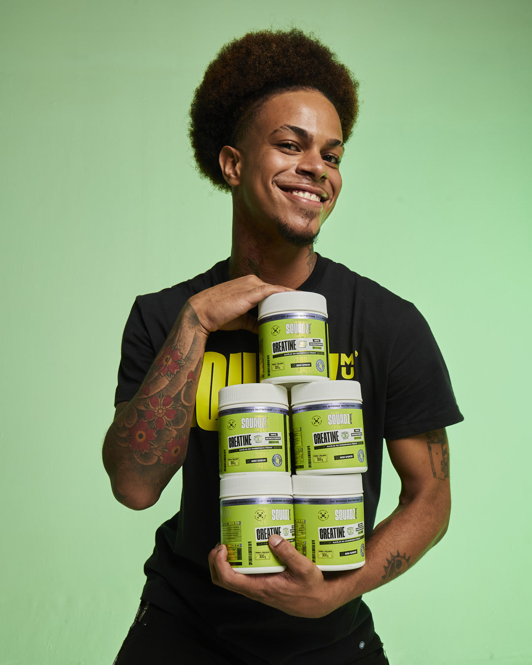 Homem equilibrando uma pilha de potes de Creatine SQUADZ MUSCLE sobre fundo verde