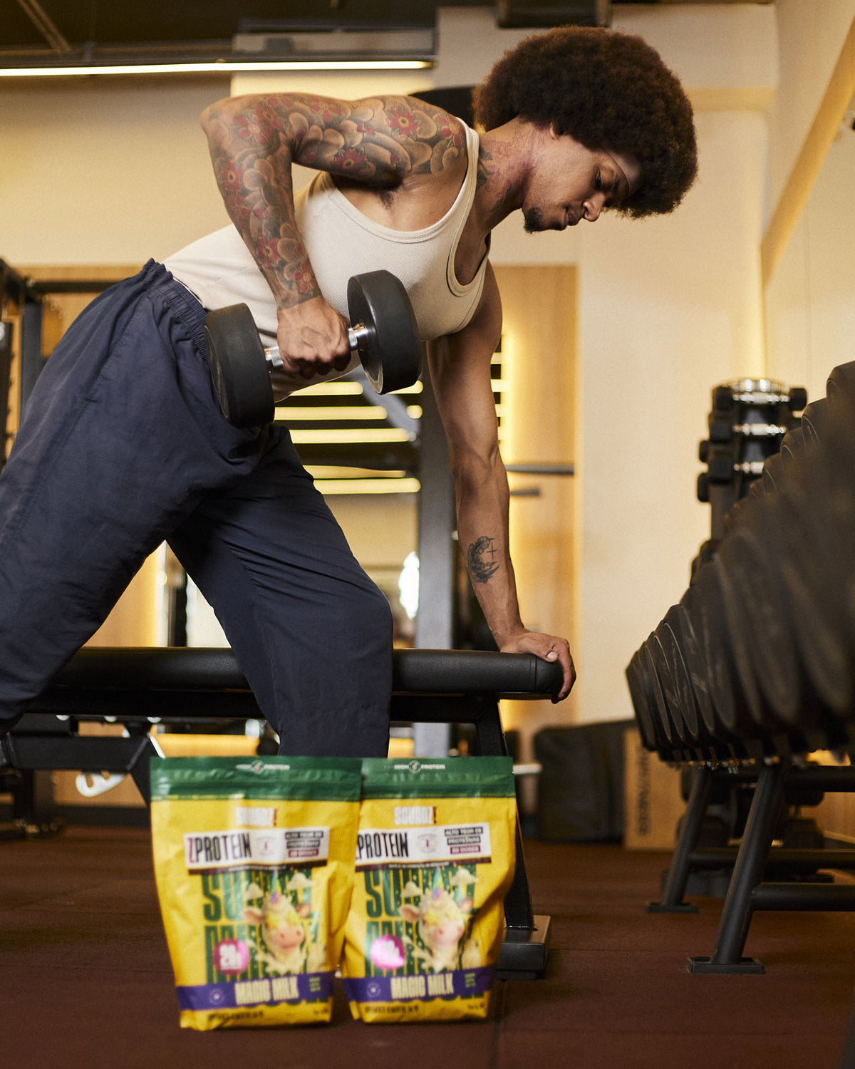 Homem treinando com halter na academia, com sacos de ZPROTEIN Leite em pó no chão